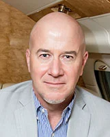 Close-up portrait of a bald man in a light jacket, seated inside a car, looking at the camera