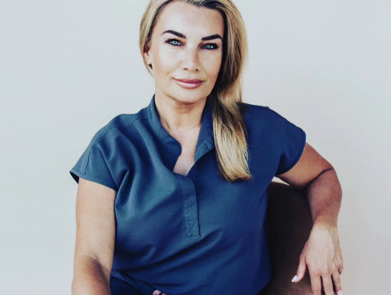 Woman with blonde hair, blue top, smiling, seated in a chair.