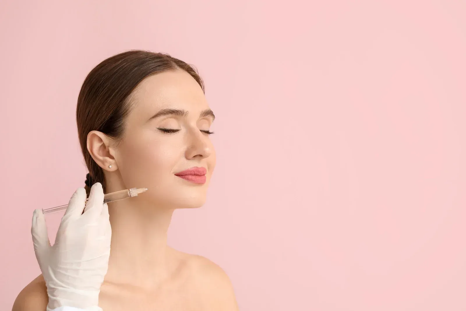 Woman receiving facial injection, white gloves, pink background.
