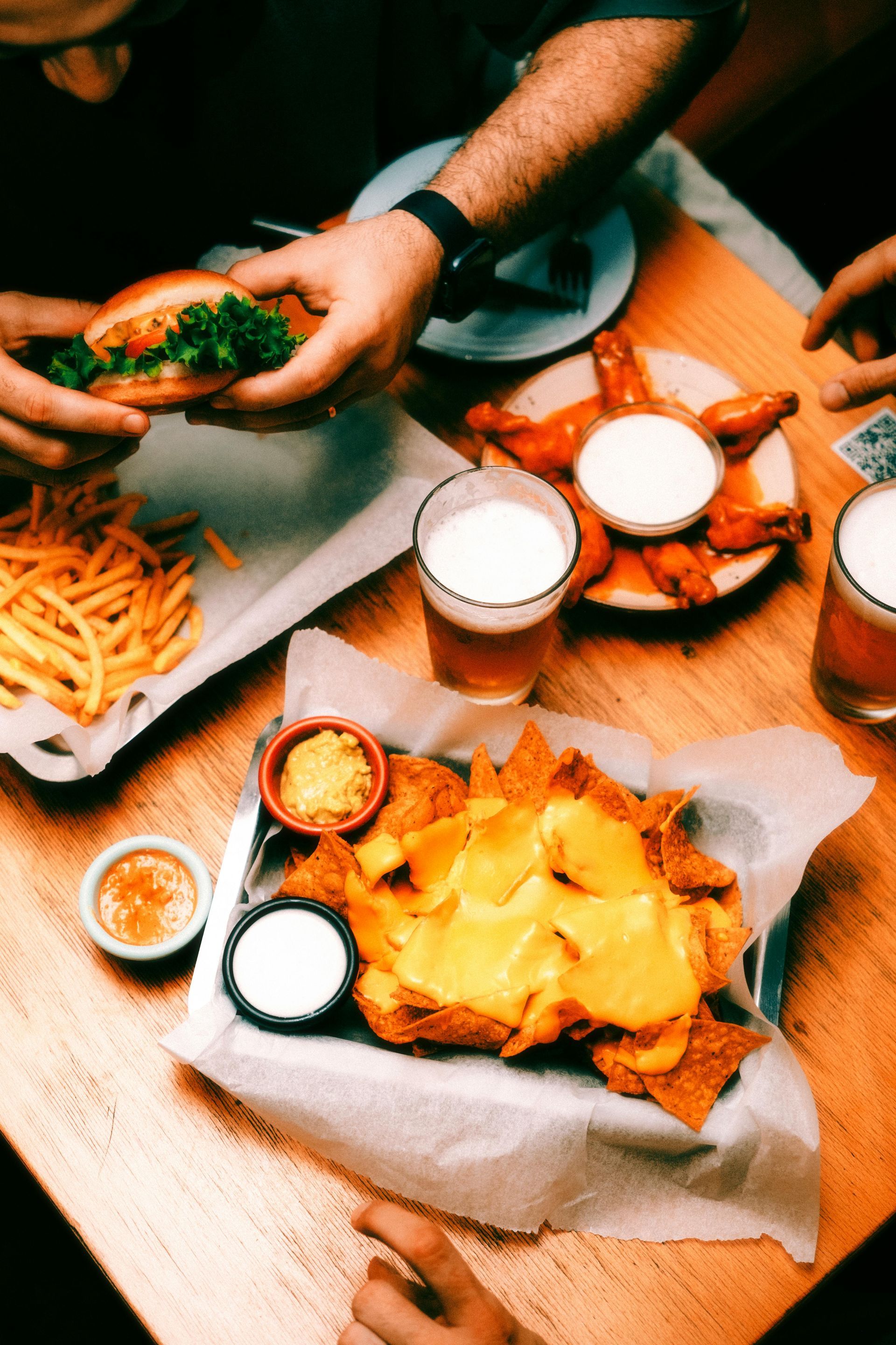 People sharing food: burger, fries, nachos, wings, and beer on a wooden table.