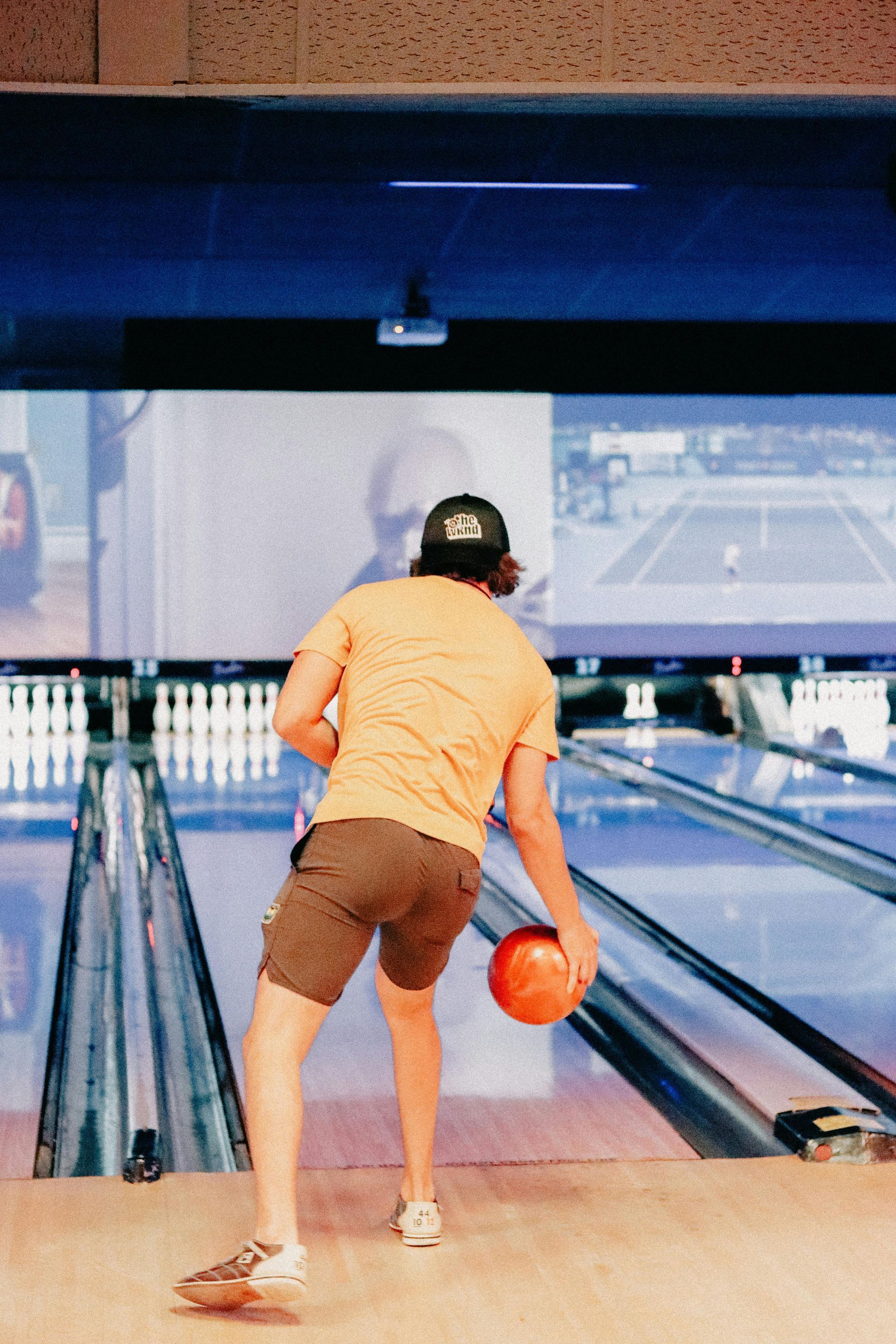 Man in yellow shirt bowling, orange ball in motion, pins visible, bowling alley setting.