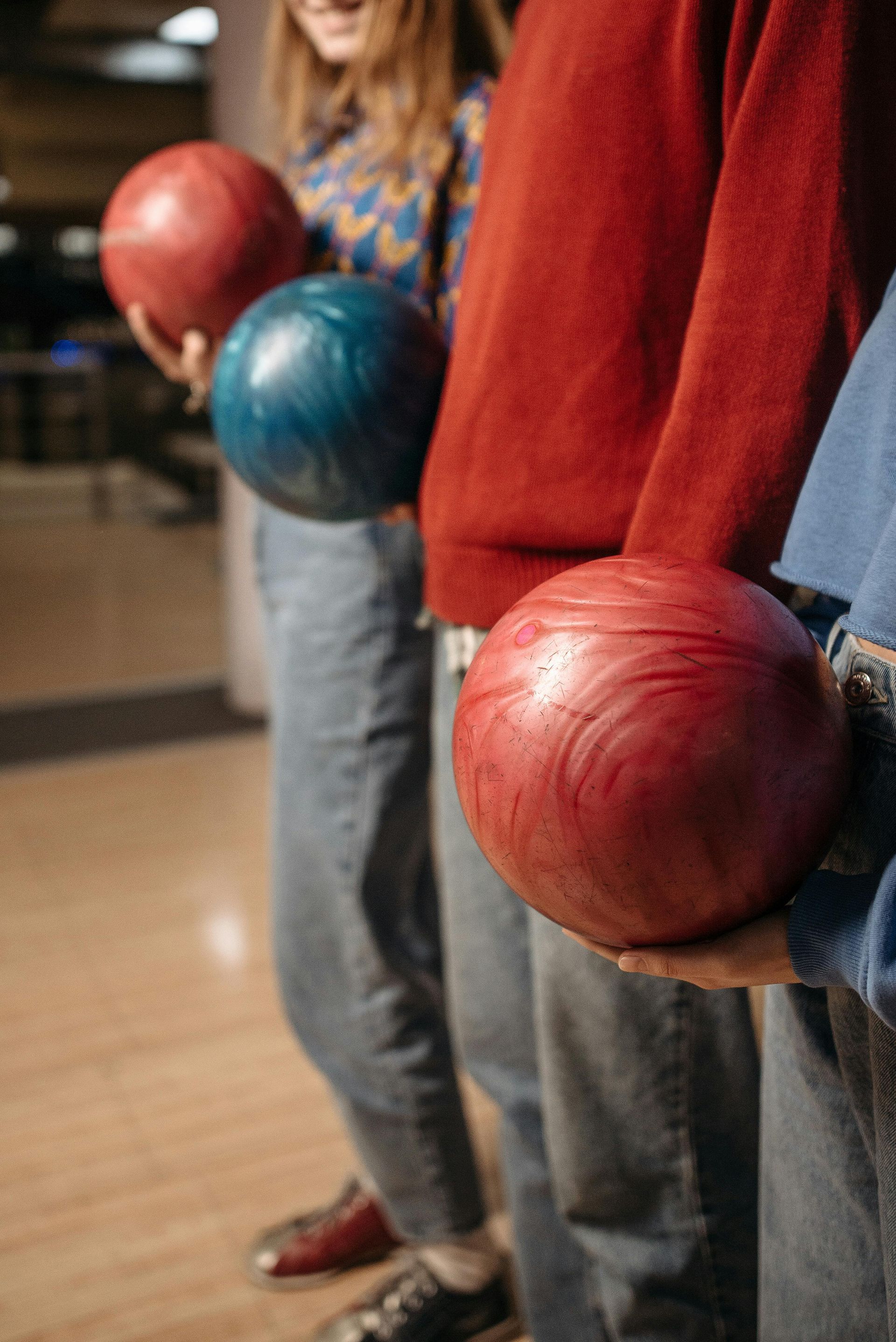 People holding bowling balls at a bowling alley. Red, blue bowling balls. Wooden floor.