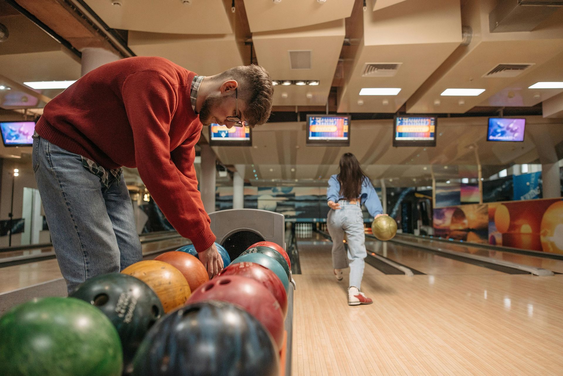 Man selecting a bowling ball as a woman approaches the lane. Indoor bowling alley setting.