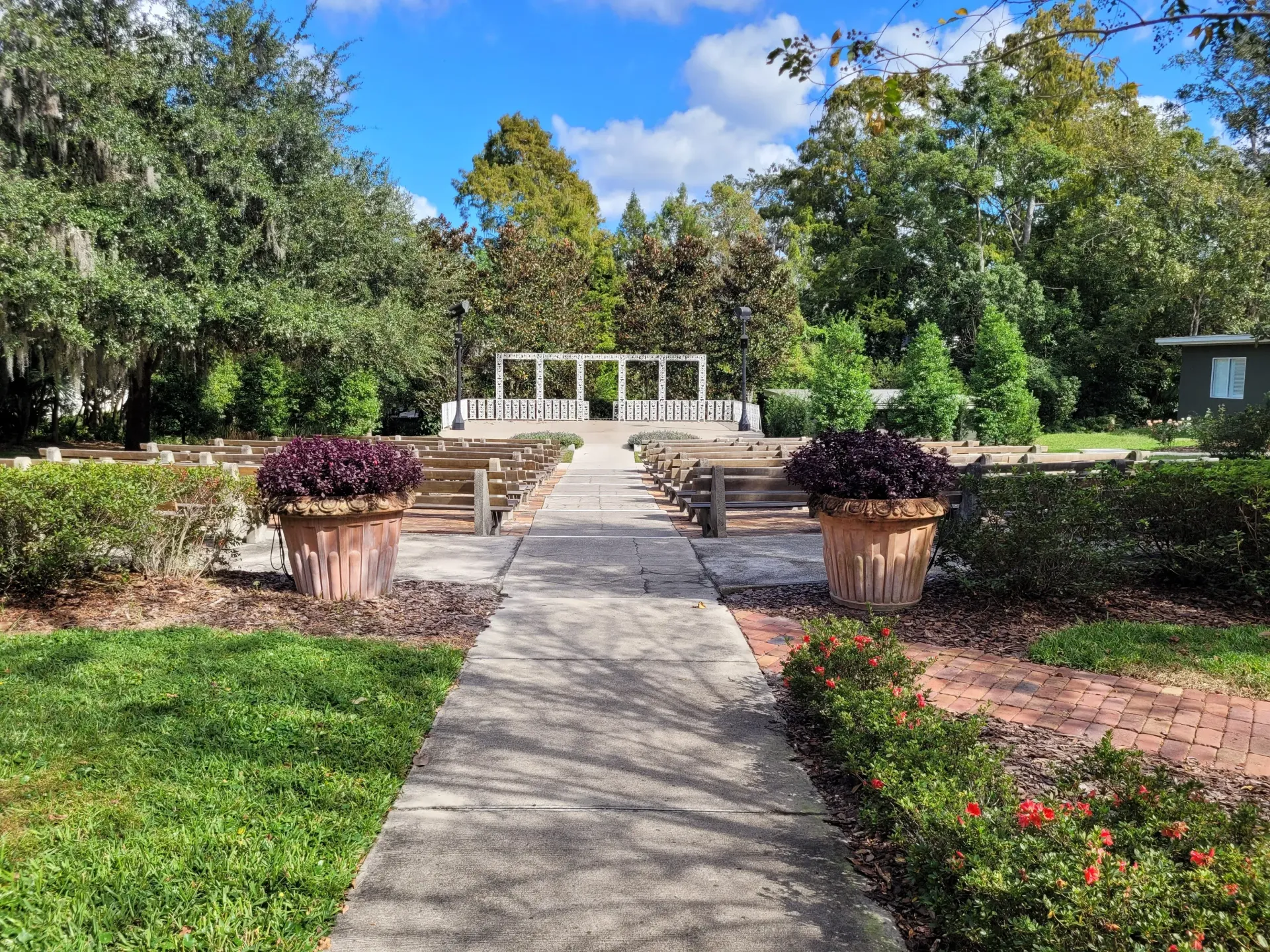 Outdoor ceremony space, stone path, wooden benches, floral pots, and green trees.