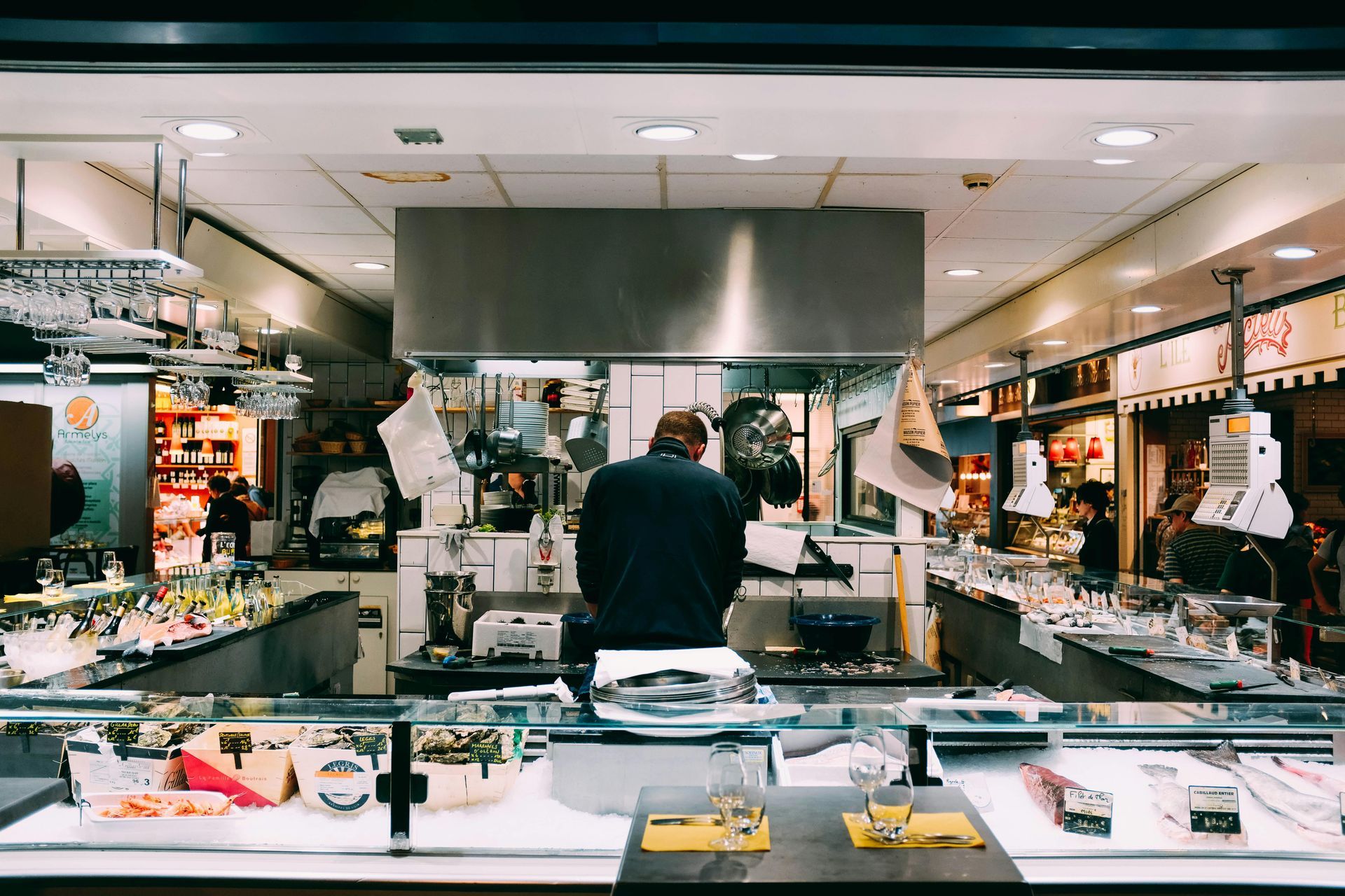 A person stands in a busy kitchen, preparing food at the counter, surrounded by display cases.