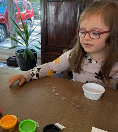 Girl with Down syndrome wearing glasses counts coins on a table. Play dough and plant are also visible.