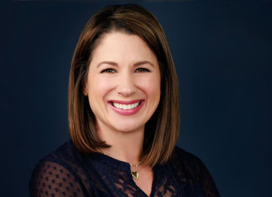 Woman with short brown hair smiles, wearing a navy top, against a dark blue background.