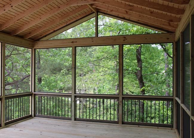 A wooden screened-in porch with a gabled ceiling, black railings, and a view of lush green trees outside.