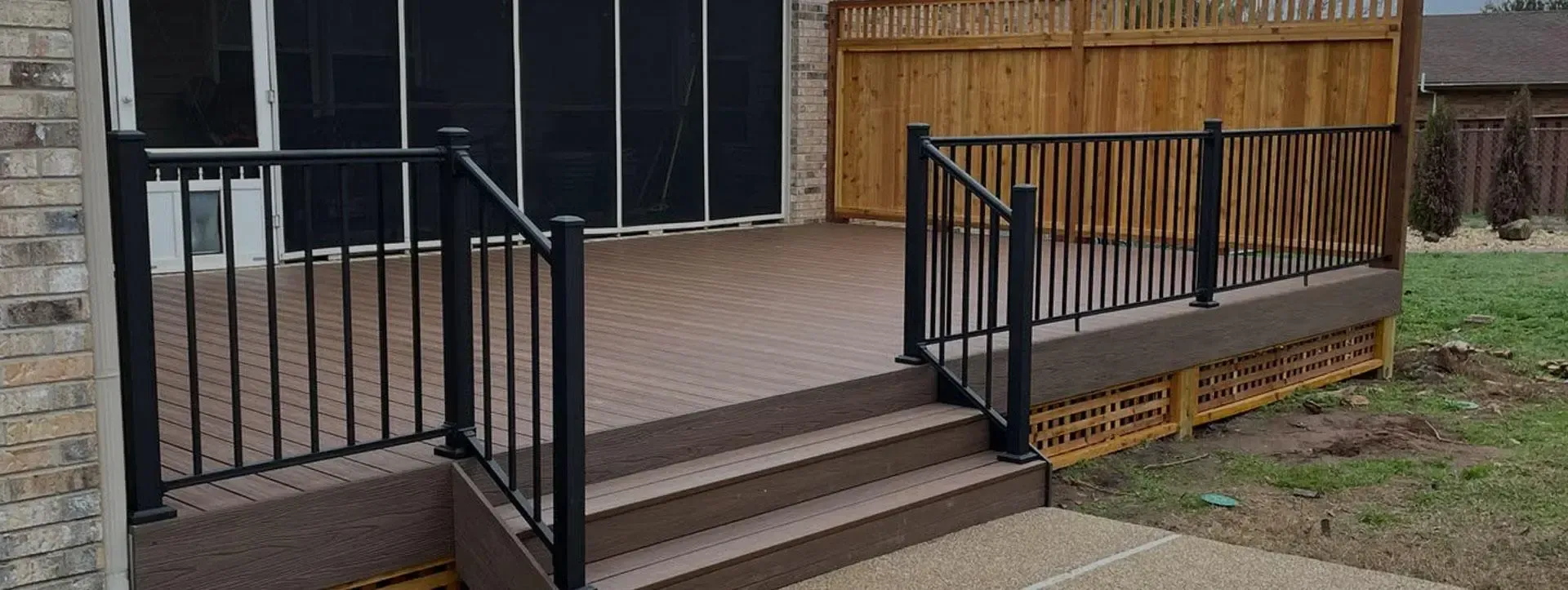 A brown composite deck with black metal railings and stairs leading to a concrete patio outside a brick house.