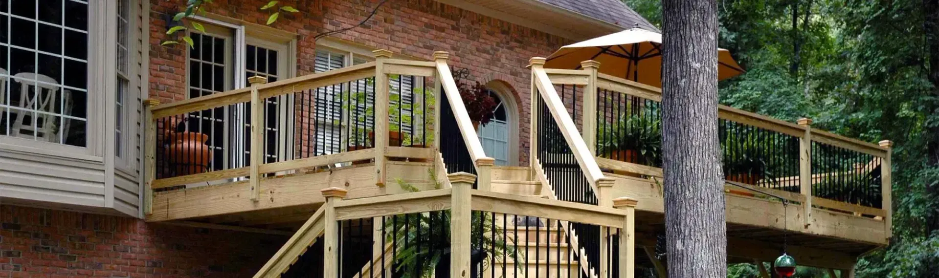 A wooden deck with railings and stairs attached to a brick house, partially obscured by a tree and an outdoor umbrella.