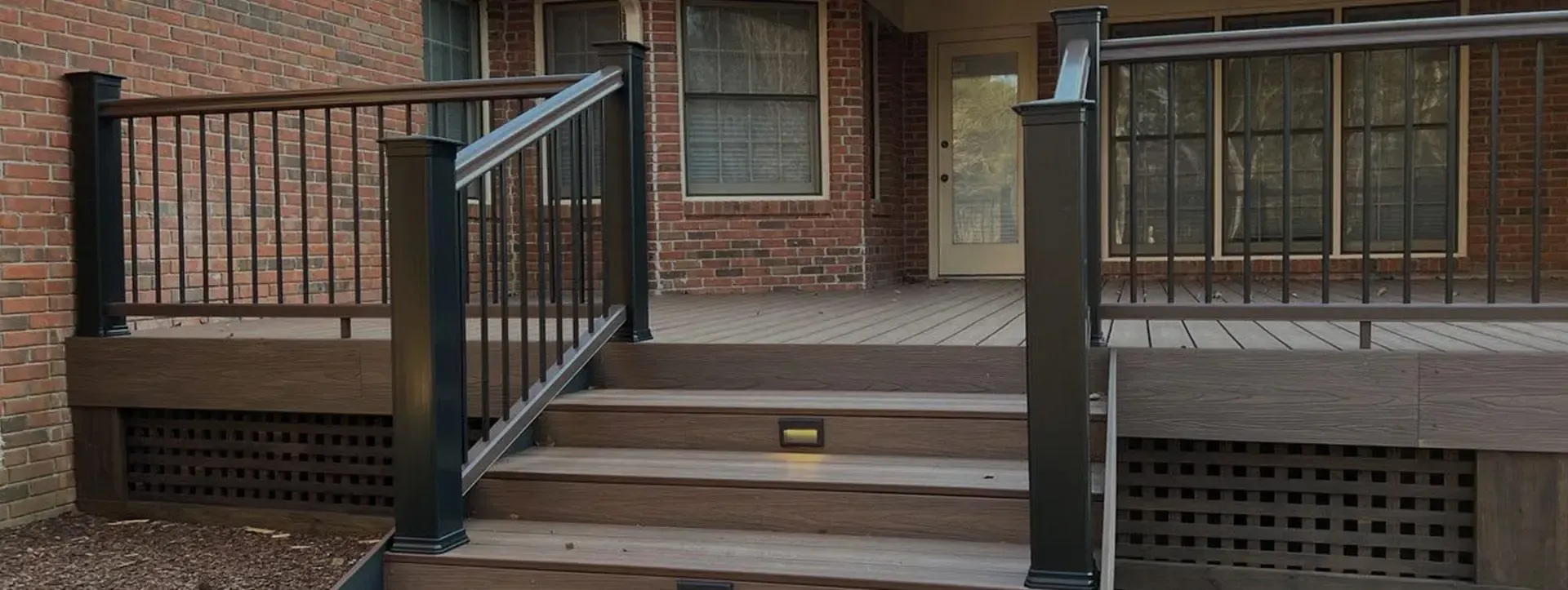 Brown composite deck with black metal railings and stairs featuring built-in lighting in front of a brick house.