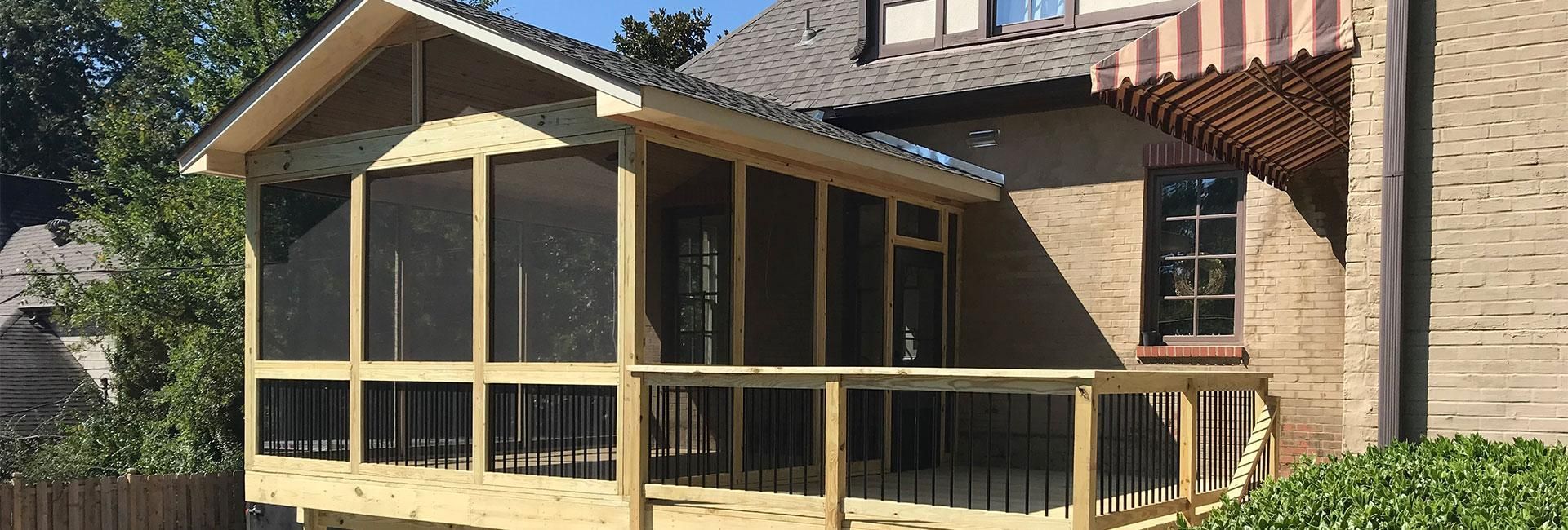 A wooden screened-in porch with a gabled roof attached to the side of a brick house with a deck and railing.