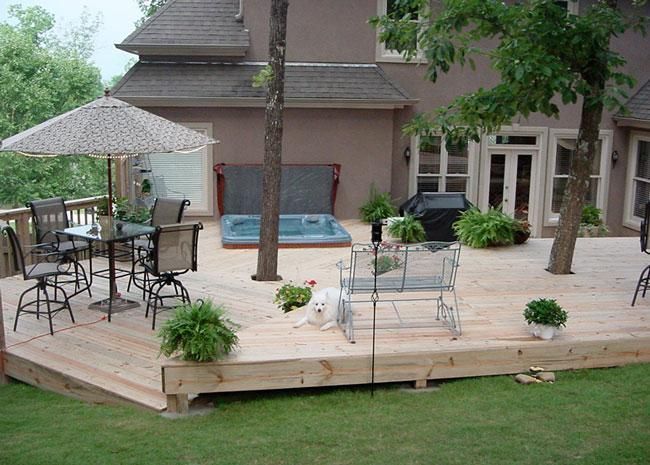 A wooden backyard deck with a hot tub, umbrella, patio furniture, potted plants, and a small white dog.