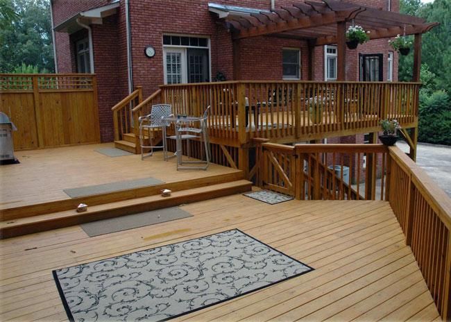 A multi-level wooden deck attached to a brick house, featuring stairs, railings, a pergola, and outdoor furniture.