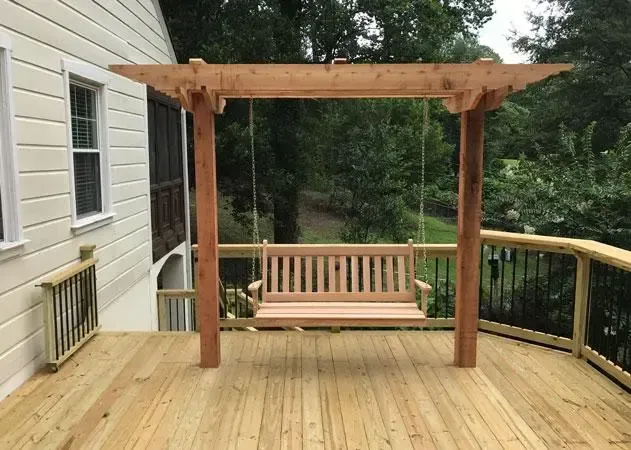 A wooden porch swing hanging from a pergola structure on a backyard deck next to a house.