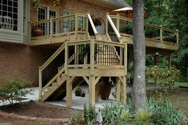 A wooden deck attached to a brick house with stairs leading down to a concrete patio in a wooded backyard.