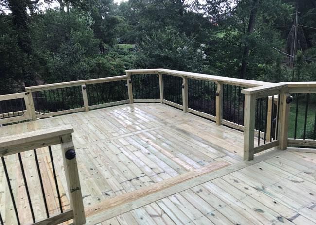A high-angle view of a new wooden deck with railings, black metal balusters, and a lush, wooded background.