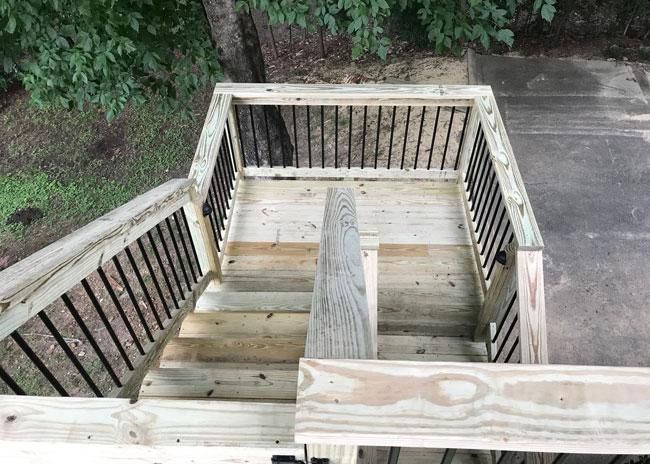 A high-angle view of a wooden staircase and landing with black metal railings, leading down to a concrete patio.