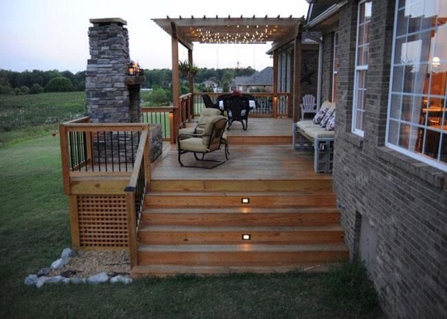 A wooden deck at dusk, featuring a stone fireplace, pergola with string lights, comfortable seating, and lit stairs.