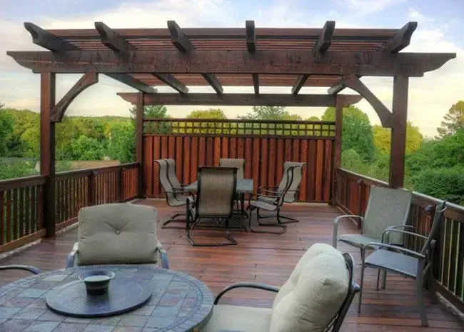 A wooden pergola on a deck with outdoor dining furniture, including a table and chairs, set against a backdrop of trees.
