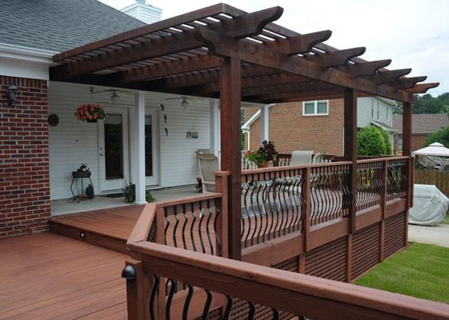 A wooden deck with a matching pergola attached to a brick house, featuring metal railings and lattice skirting.