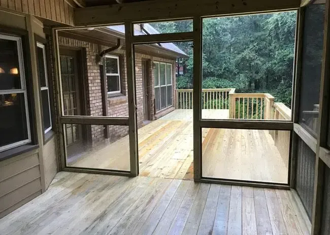 A screened-in porch with light wood flooring overlooking an outdoor wooden deck surrounded by trees.
