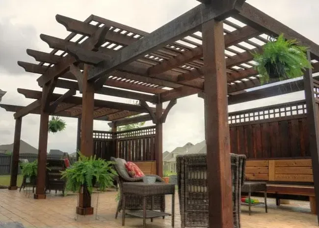A dark wood pergola on a patio with wicker furniture, hanging plants, and a privacy screen.