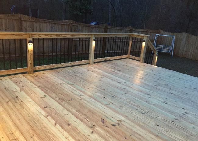 A wooden deck featuring a railing with black metal balusters and built-in post lights at twilight.