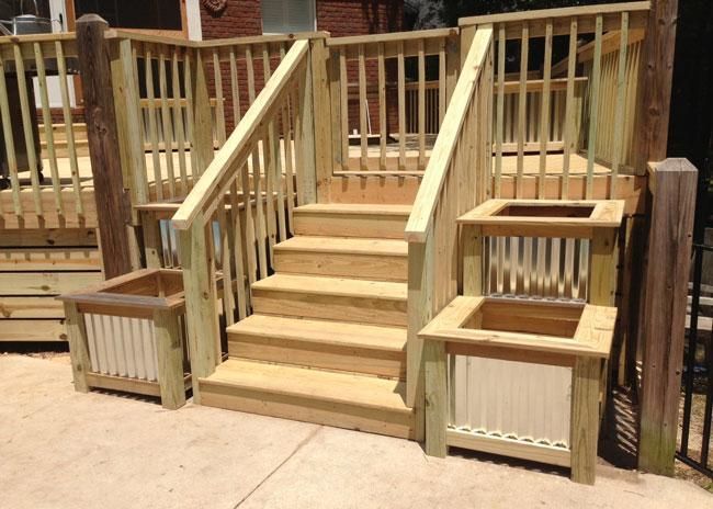 Wooden deck stairs featuring railing and two planters with corrugated metal siding on a light concrete patio.