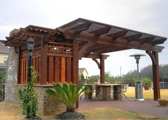 A wooden pergola with a stone wall base, outdoor heater, and seating area on a paved patio in a suburban backyard.