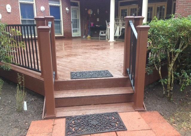 A set of brown wooden stairs leading up to a large, elevated deck with black metal railings in front of a brick house.