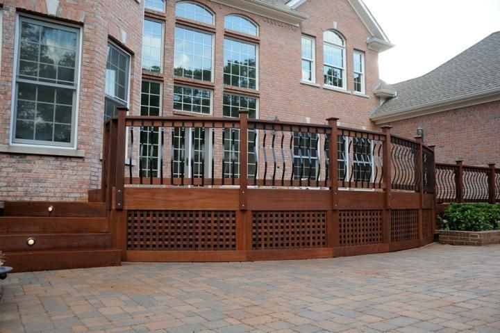 A wooden deck with lattice skirting and black metal railings sits against a brick house above a paver patio.