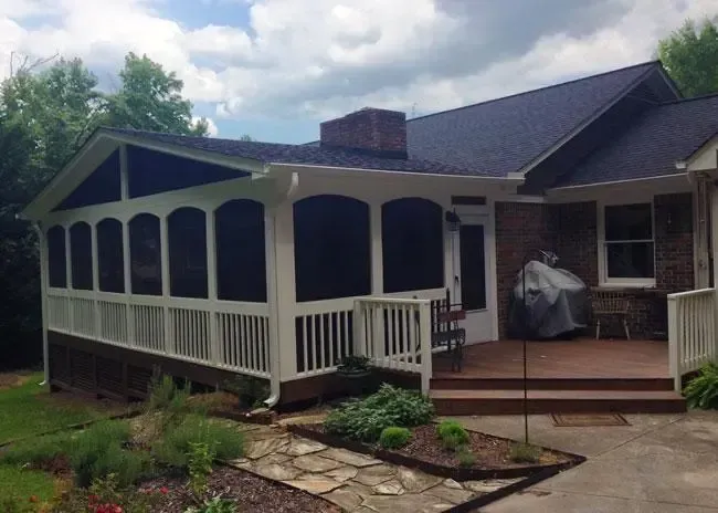 A covered screened-in porch with a white railing and frame, adjacent to a brick house with a wooden deck and a stone path.
