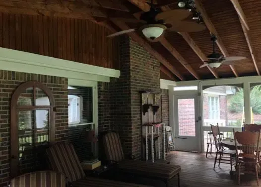 A screened-in porch with a brick wall, wooden ceiling beams, two ceiling fans, lounge chairs, and a dining set.