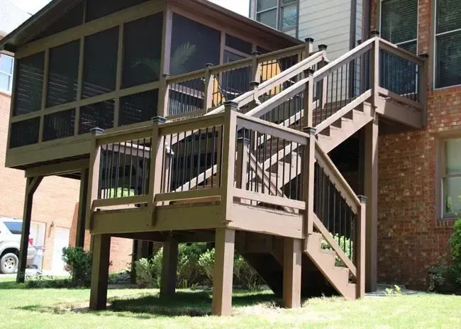 A brown wooden deck with a screened-in porch and stairs leading down to a grassy yard next to a brick house.
