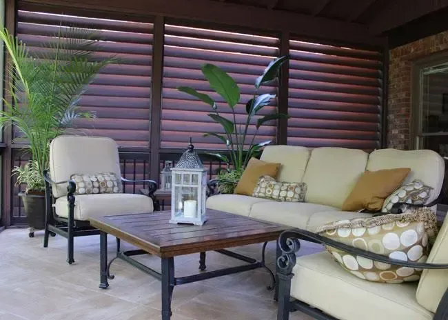 A patio with light-cushioned furniture, a wooden coffee table, a lantern, and potted plants in front of wooden shutters.