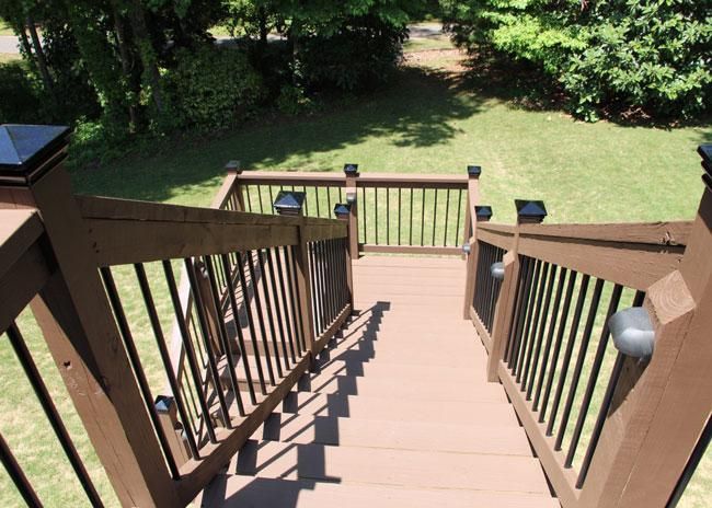 A downward view of a brown wooden deck staircase with black railings leading to a grassy backyard.