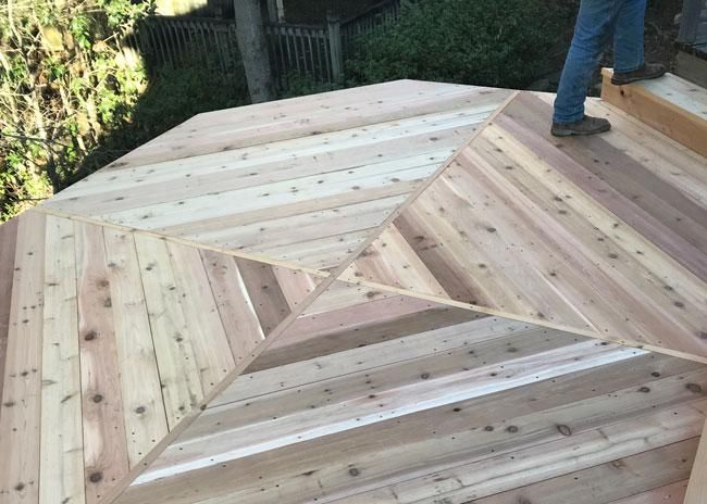 A person stands on a freshly built wooden deck featuring a geometric, multi-directional pattern of light cedar planks.