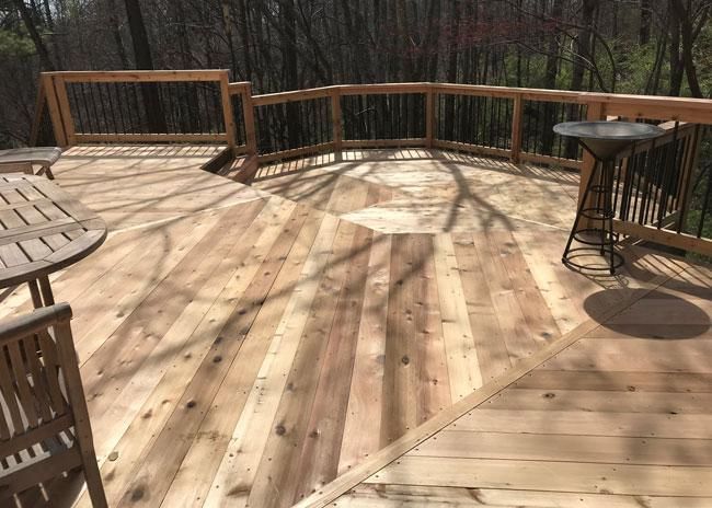 A wooden deck with a geometric pattern, railing, a round table, and a birdbath overlooking a wooded area.