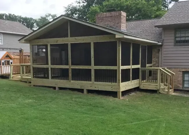 A wooden screened-in porch with a peaked roof attached to the back of a brick and siding residential home.