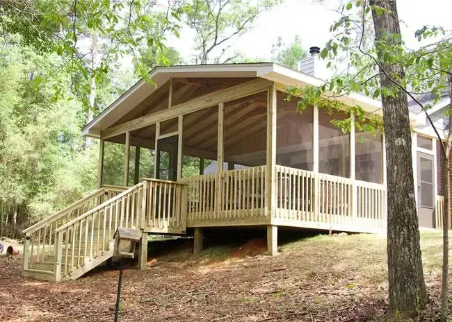A wooden screened-in porch with a staircase leading up, surrounded by trees in a wooded yard.