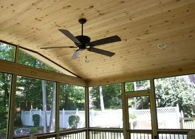 A screened-in porch with a wooden ceiling, a dark ceiling fan, and views of a backyard and trees.