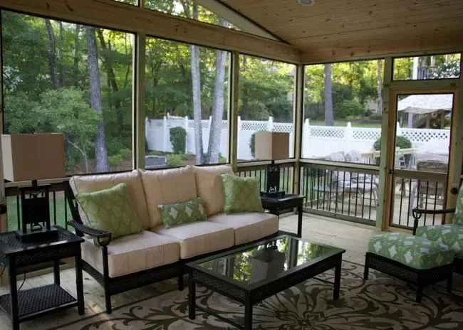 A screened-in porch with a beige sofa, green patterned cushions, a glass coffee table, and a view of a backyard and fence.