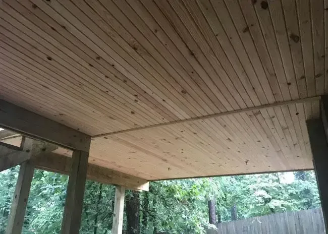 A wooden tongue-and-groove ceiling under a porch roof, looking out toward trees and a fence.