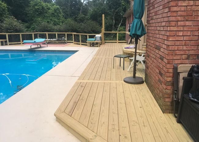 A wooden pool deck beside a blue pool and a red brick house wall, featuring patio furniture and an umbrella.