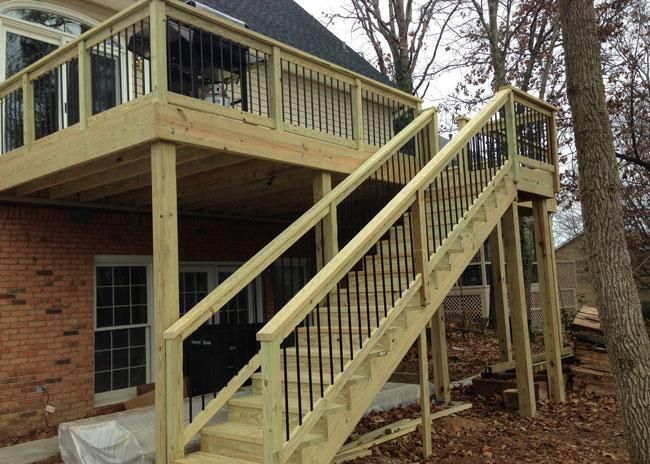 A raised wooden deck with black metal railings and a staircase leading down to the ground beside a brick house.