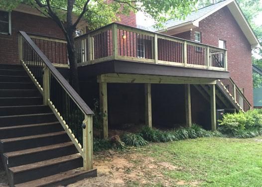 A wooden deck with a staircase leading up to a brick house exterior, surrounded by a lawn and a tree.