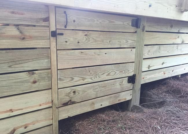 A horizontal wooden deck skirting panel with a hinged access door, featuring a black handle, latch, and two hinges.