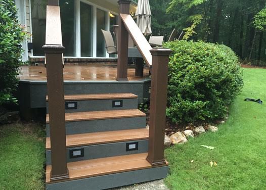 Wooden steps with built-in lights lead up to a gray deck in a backyard with a green lawn and bushes.