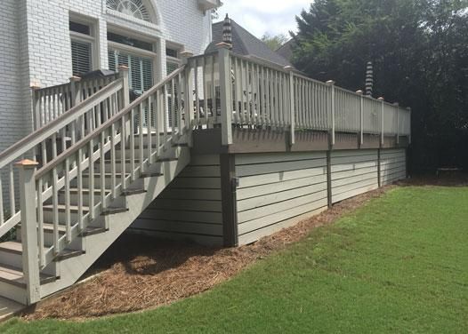 A white house with a raised wooden deck, staircase, and horizontal siding underneath, situated next to a green lawn.